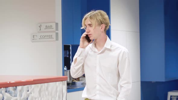 Portrait of Serious Millennial Blond Man Talking on the Phone Standing in Office Corridor