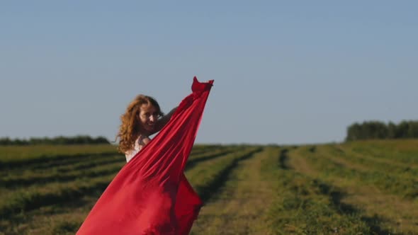 Redhead Woman in White Dress with Red Cloth in Her Hands on Background of Field