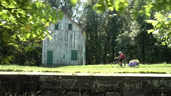 Mature woman pulling a trailer with a dog in it cycles past an old white wooden building on the C&O