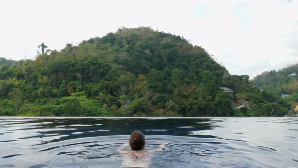 Lady Swims to Pool Edge Against Picturesque Hill with Forest