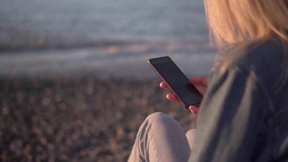 blonde in denim suit is sitting on seashore at sunset with smartphone in hands