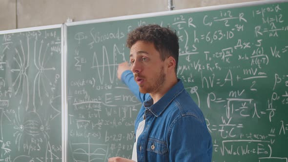 Caucasian Young Male Teacher Standing at Blackboard with Formulas Graphics and Laws and Explaining