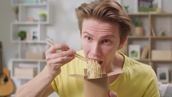 Young Man Eating Noodles From Box with Chopsticks at Home in the Living Room