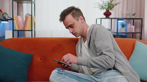 Man Sitting on Couch Using Credit Bank Card and Smartphone While Transferring Money Online Shopping