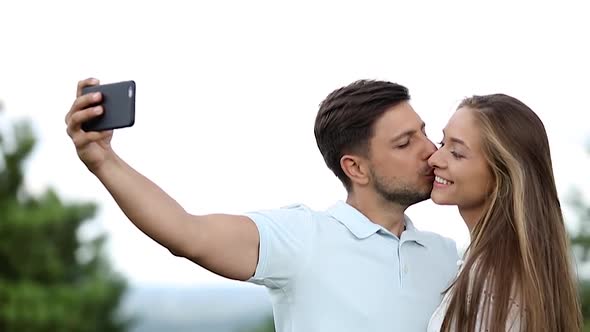 Man Kissing Woman And Making Photo On Mobile Phone In Nature