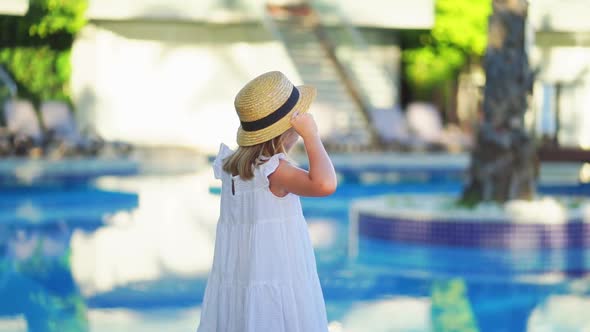 Cute Little Girl in a White Dress and Straw Hat By the Pool Near the Villa