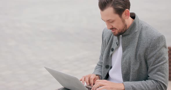 Young Man with Mustaches and a Beard Sitting on a Bench in the Square Typing on the Laptop Keyboard