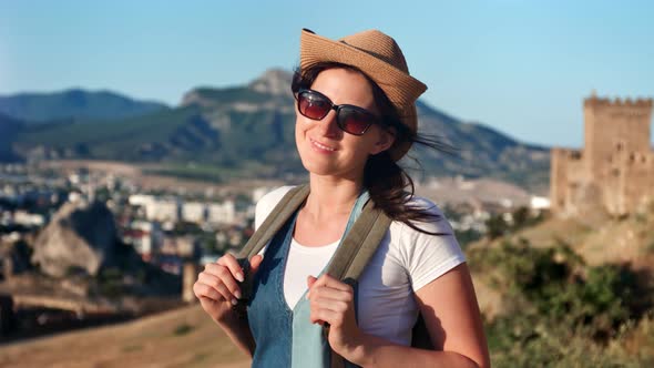 Smiling Travel Woman Wearing Sunglasses and Hat Posing at City Surrounded By Mountain Landscape