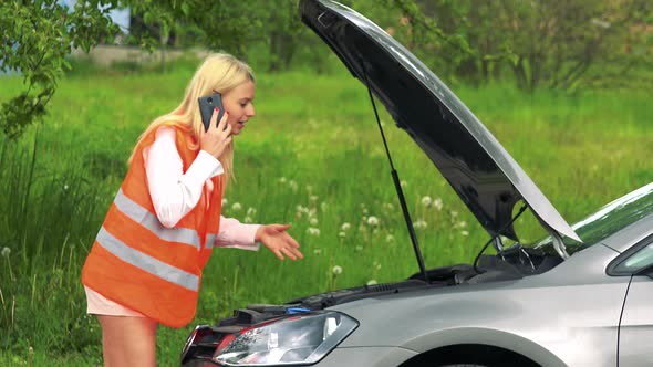 Young Attractive Blond Woman Wears Warning Vest and Phones with Smartphone in Front of Broken Car