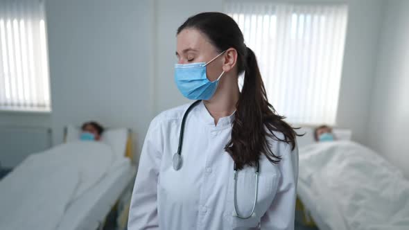 Portrait of Exhausted Overworked Nurse Looking Back at Patients in Beds Wiping Forehead Looking at
