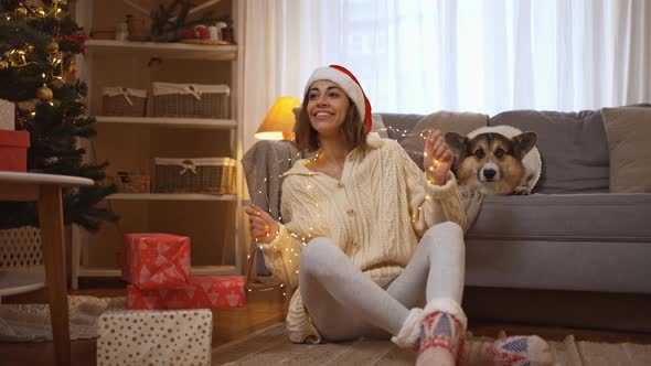 Emotional Smiling Young Woman in Santa Hat Holding Lights and Her Funny Corgi Dog Sits at Couch in