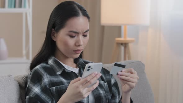 Angry Asian Woman Sit on Sofa at Home Hold Credit Card Use Mobile Phone Try to Pay with Ebank App
