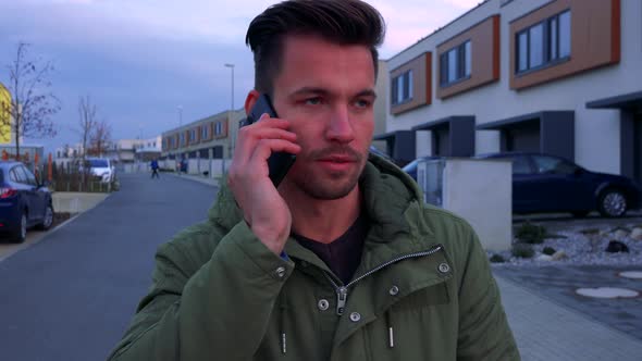 A Young, Handsome Man Stands on a Road in a Neighborhood and Talks on a Smartphone