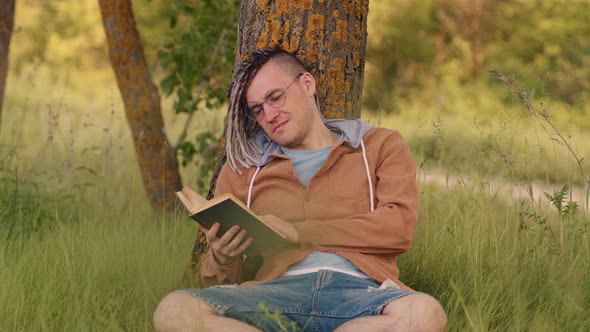 A man in casual clothes and glasses reads literature under a tree.