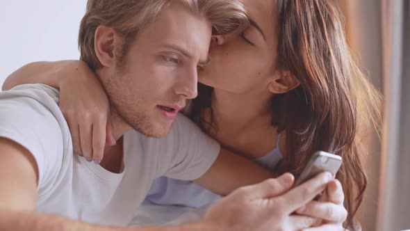 Beautiful Caucasian Female Kissing Her Boyfriend While He is Concentrated on Mobile Phone Sitting in