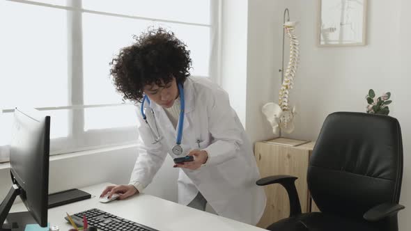 Female Doctor Using Mobile Phone While Working on Laptop in the Consultation