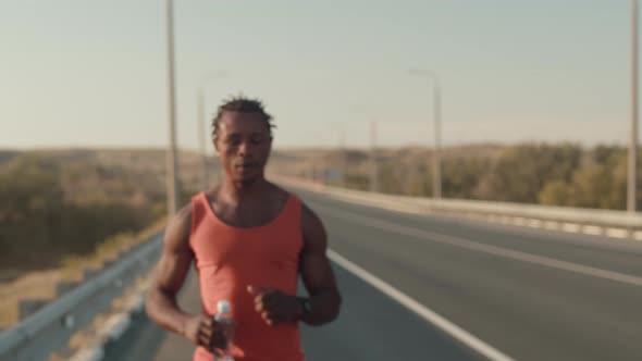 Black man runs along road doing running workout, slowly stops and drinks water from bottle