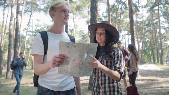 Portrait of Positive Man and Woman Checking Route Using Paper Map As Blurred Friends Making Photos