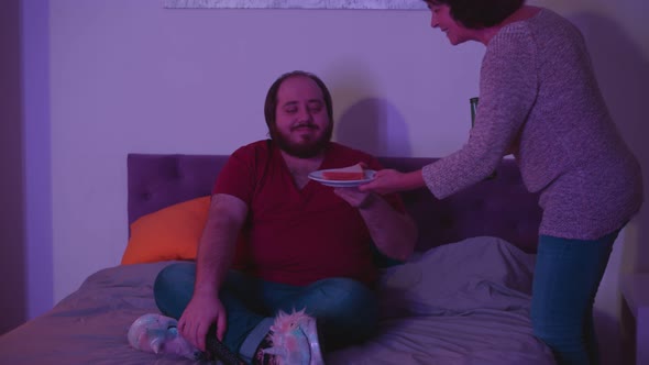 Young Fat Guy Watching Tv in Bed While Senior Mother Bringing Him Beer and Sandwich