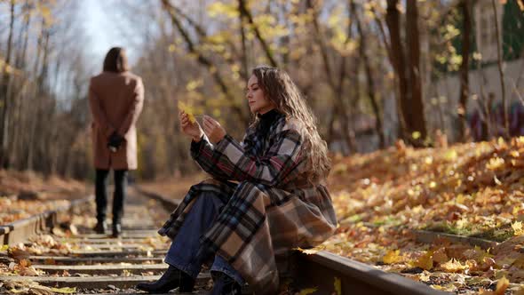 Woman is Sitting on Tram Rail in Autumn Day Playing with Leaf Silhouette of Man in Background