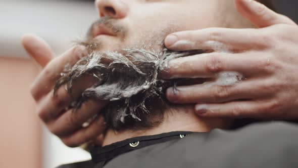 Male barber's hands wash beard of a Caucasian man with shampoo, close-up. Professional hands of the