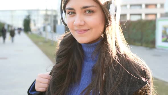 Young woman portrait in the city back light listening music headphones looking camera smiling