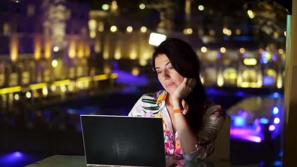 Beautiful Young Brunette Woman Uses Laptop, Sitting at Table on Open Terrace or Balcony, Against