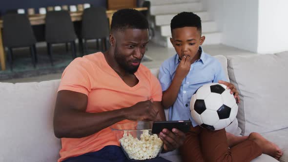 African american father and son eating popcorn and watching a football game on smartphone