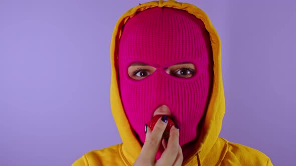 Close Up of Young Woman in Pink Balaclava and Yellow Hood Eats Strawberry on Purple Background