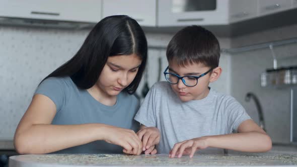 Cheerful Boy and Girl Will Put Together a Puzzle While Sitting at a Table at Home in the Kitchen