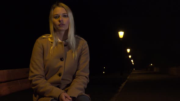 A Young Beautiful Woman Sits on a Bench and Looks Seriously at the Camera in an Urban Area at Night