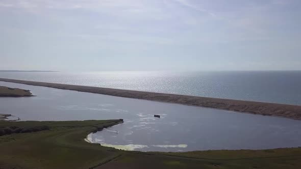 Wide aerial tracking forward to the fleet lagoon at the west end of the shingle beach. Sun reflectio