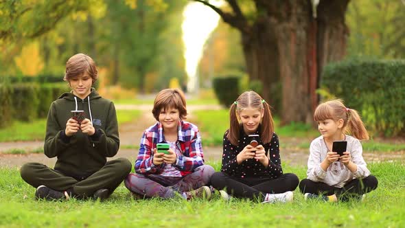 Happy Children Friend Girls Group Sit with Cross Legs on Grass and Playing Internet Game with Mobile