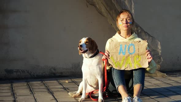 Ukrainian Refugee Woman with National Flag on Cheek Sitting Underground with Dog and Sign No War