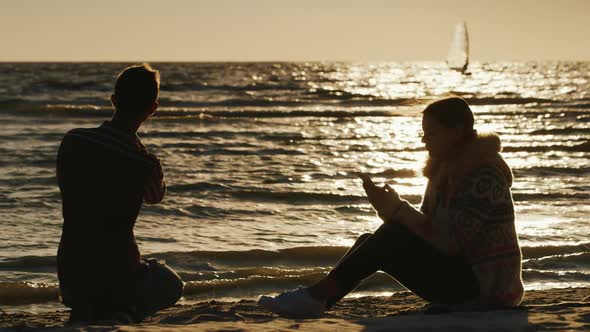 Romantic Couple Relaxing on the Beach at Sunset. Woman Enjoying a Telephone, a Man Throws Stones