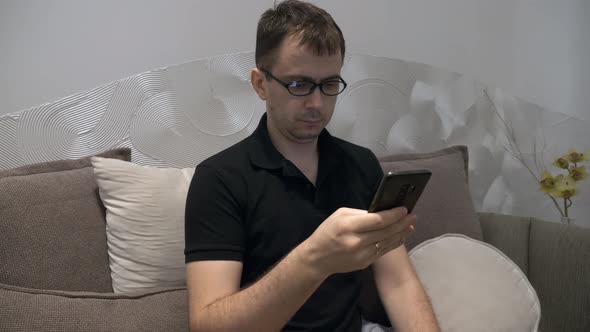 Serious Man in Black Polo Tshirt and Glasses Sitting on Couch with Mobile Phone