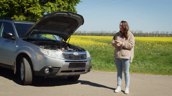 Woman Standing at Broken Car Searching for Repair Service on Smartphone