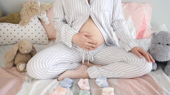 Pregnancy Concept. Pregnant Woman Choosing Socks For Future Baby Sitting In Bed In Nursery.
