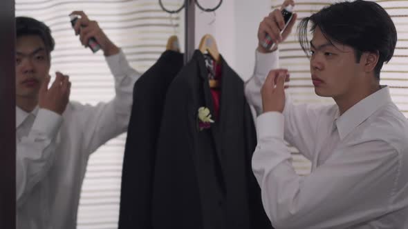 Serious Asian Young Groom Spraying Hair Spray Looking in Mirror in Bathroom