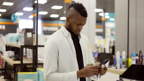 Concentrated Man in Mask is Choosing a New Mobile Phone in a Shop Checking How It Works