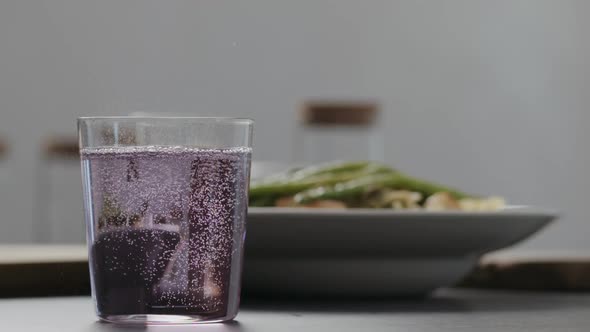 Slow Motion Purple Fizzy Drink in a Tumbler Glass on Concrete Countertop with Pasta on Background