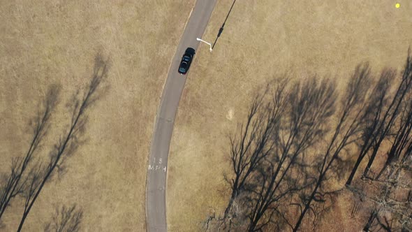 A top down shot over a single car driving through an empty park on a sunny day. The drone follows th
