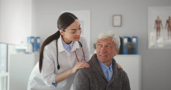 Female Doctor Hugging Smiling Senior Patient and Looking at Camera in Clinic Office
