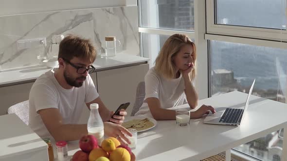 Couple's Morning They're Sitting By a Counter in Kitchen Woman Using Laptop and Man Smartphone