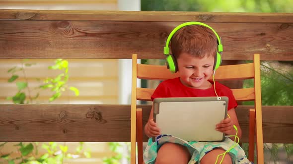 The Little Boy in Earphones Listens To Music on the Digital Tablet 