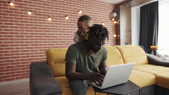 Busy Young African Father Doing Freelance Work on Laptop While His Son Disturbing Him From the Back