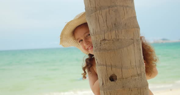 Happy Little Girl on the Beach in a Swimsuit and a Big Hat Looks Out From Under a Palm Tree Looks