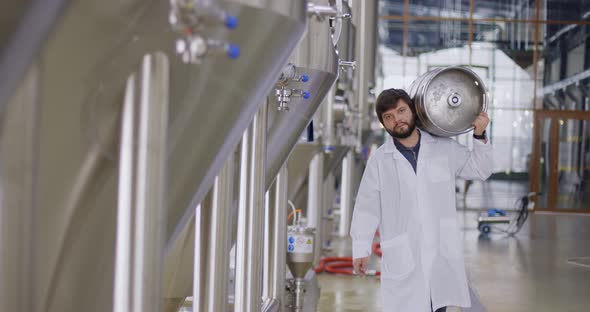 A Working Male Brewer in Lab Coat Carries a Keg on Shoulder Filled with Beer As He Passes Beer Tanks