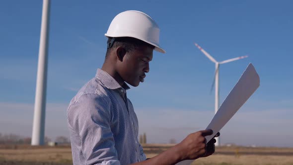 African American Electrician Engineer in a Helmet Stands Against the Backdrop of a Windmill at an