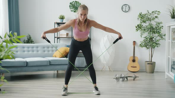 Portrait of Beautiful Blonde in Sportswear Doing Resistance Band Exercise Indoors in Modern House
